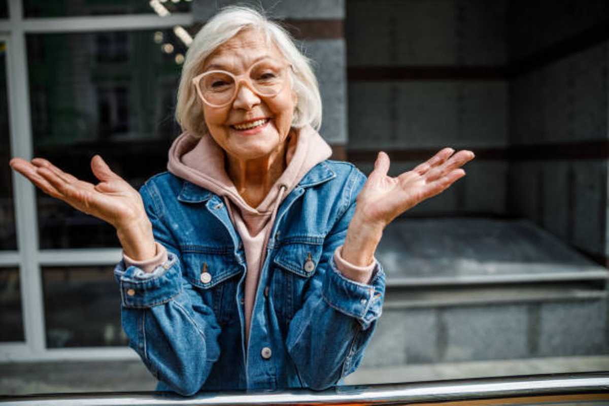 Smiling elderly woman in glasses and a denim jacket shrugging playfully with raised hands while standing outdoors.
