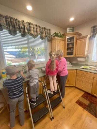 Elderly woman helps three young children wash dishes at the kitchen sink, with two kids standing on step stools.
