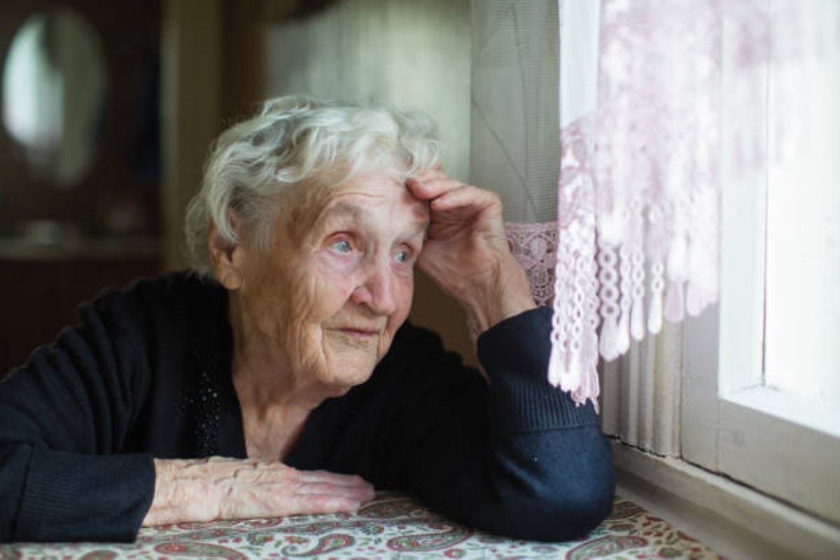 Elderly woman with white hair rests her head on her hand while gazing out a window, appearing thoughtful and serene.