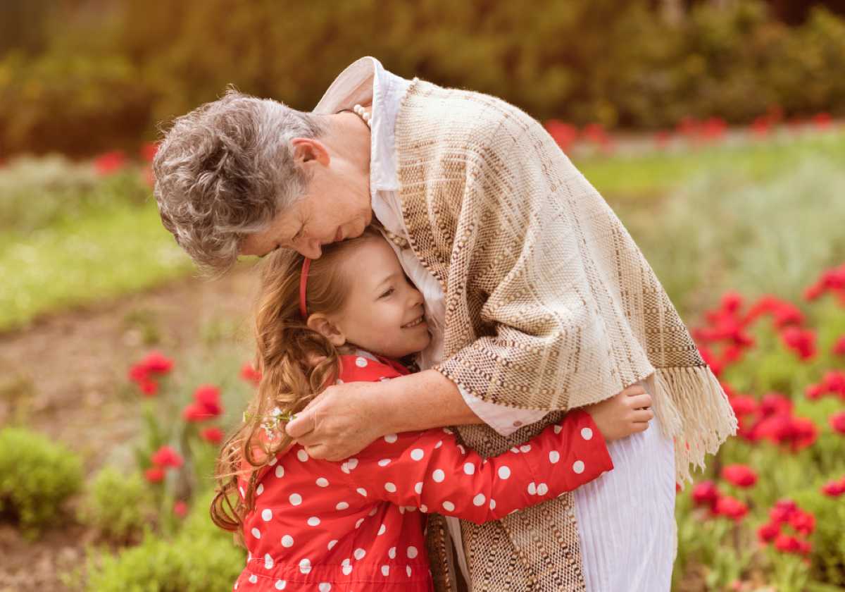 Elderly woman hugs a smiling young girl in a red polka dot coat in a garden with blooming red flowers.