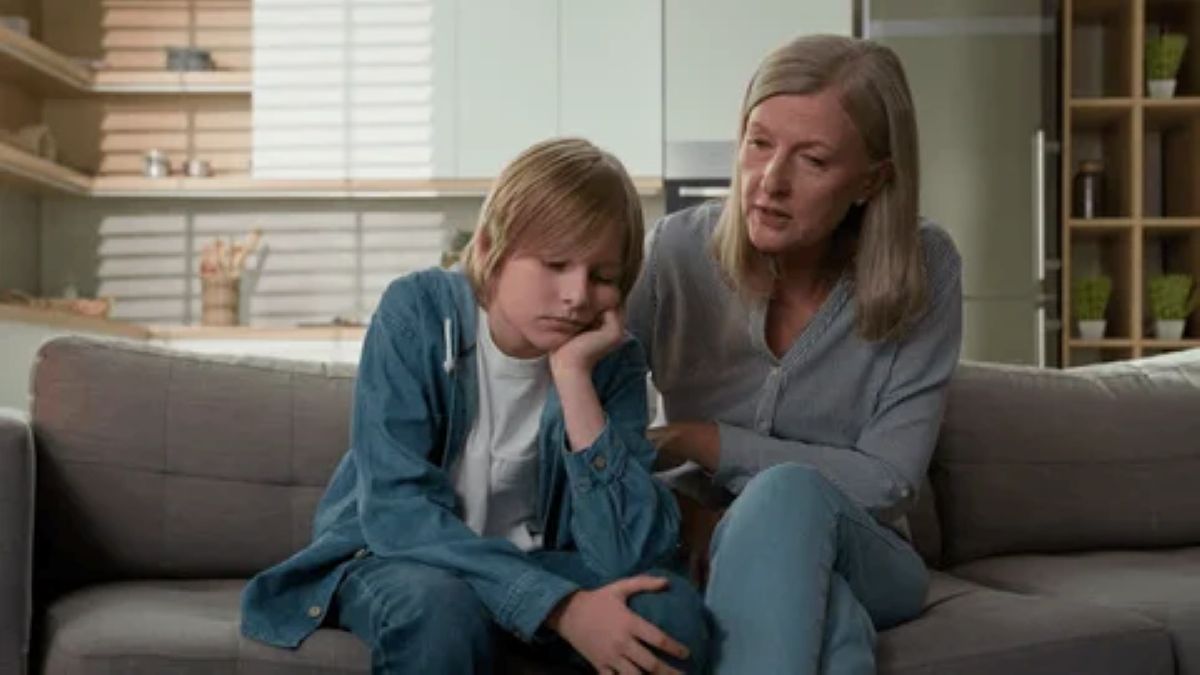 A woman and a boy sit closely on a couch, with a modern kitchen visible in the background, creating a warm, intimate atmosphere.