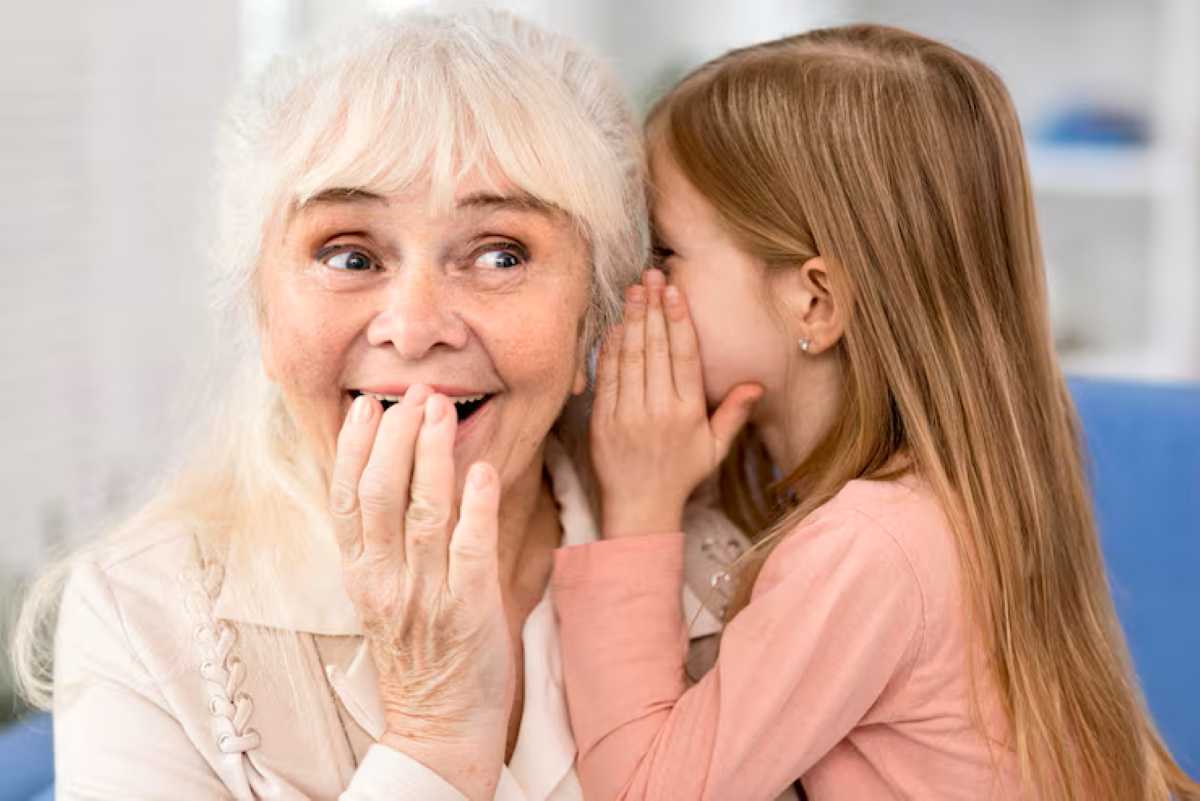 An elderly woman and a young girl share a playful moment, whispering secrets to each other in a cozy indoor setting.
