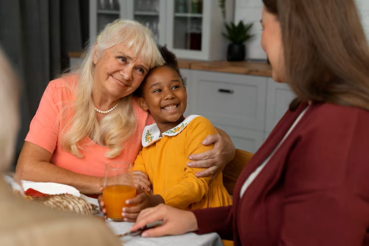 A grandmother and young girl share a warm embrace at a dining table, surrounded by food and drinks in a cozy kitchen setting.