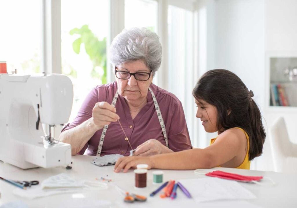 Grandmother sewing with a young girl beside her, surrounded by fabric and thread at a bright workspace.