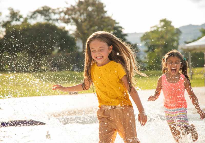 Two girls laughing and running through water spray on a sunny day in the park.