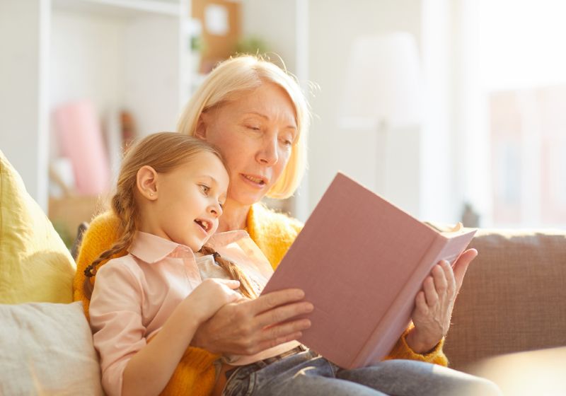 Grandmother reading a book to her granddaughter while sitting together on a cozy couch.