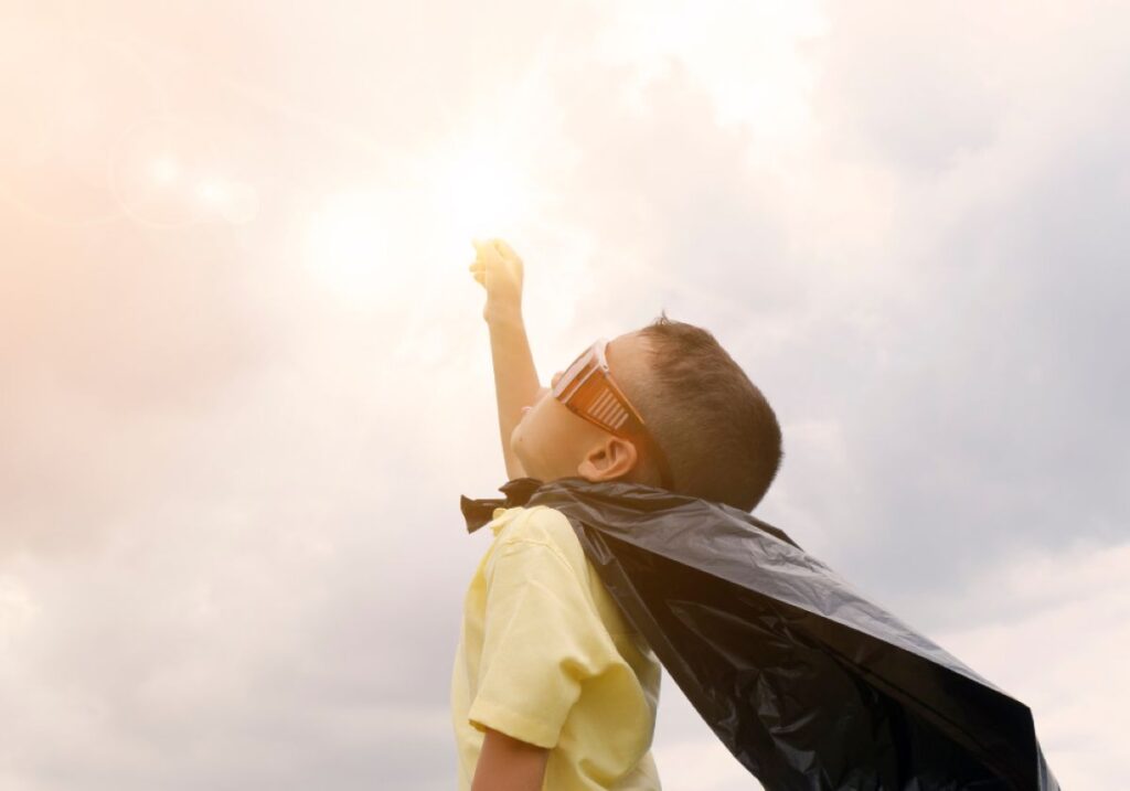 Young boy wearing a cape and sunglasses raises his fist toward the sky, pretending to be a superhero in the sunlight.