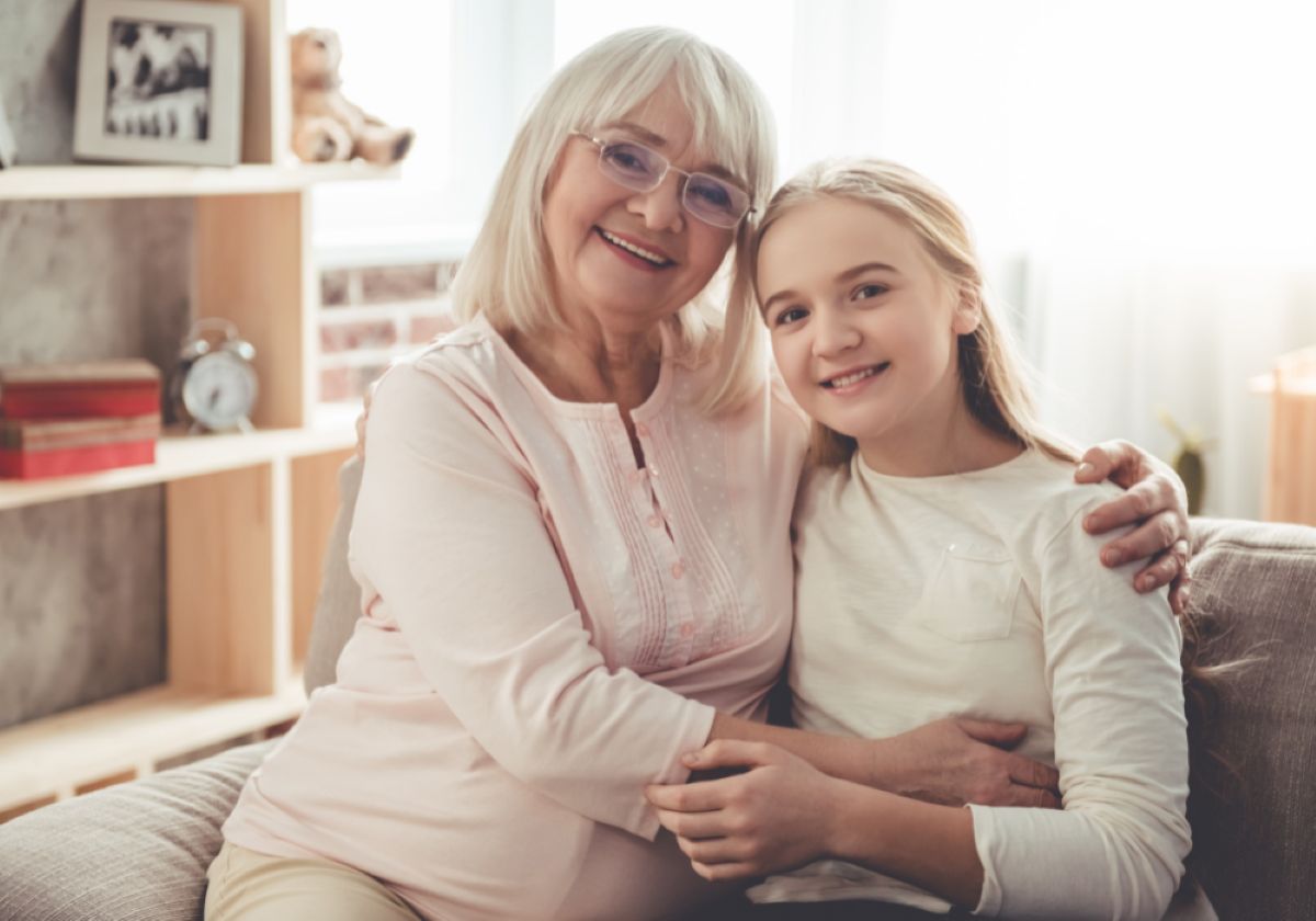 Grandmother and granddaughter sit close together, smiling warmly and hugging on a cozy living room couch.