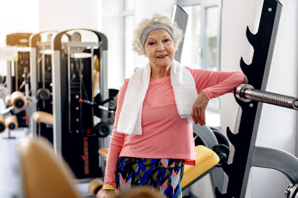 Elderly woman in workout clothes smiles confidently at the gym, with a towel around her neck near weight machines.
