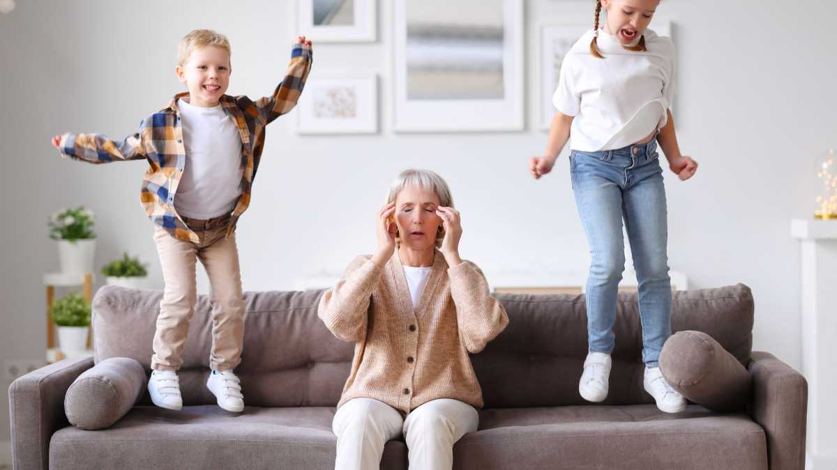 Tired grandmother sits on a couch rubbing her temples while two energetic kids jump on the cushions beside her.