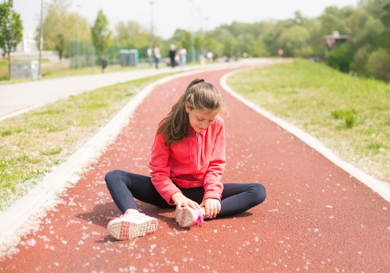 Girl in a red hoodie sits on a running track, holding her ankle with a pained expression after a possible injury.