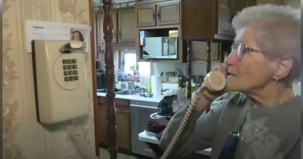 Elderly woman using a corded wall phone in a vintage-style kitchen with wooden cabinets and wallpaper.