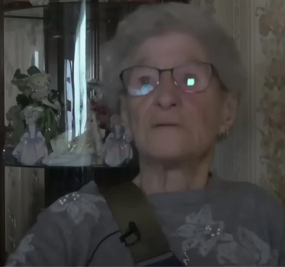 Elderly woman wearing glasses and a floral sweater sits near a display case with porcelain figurines.