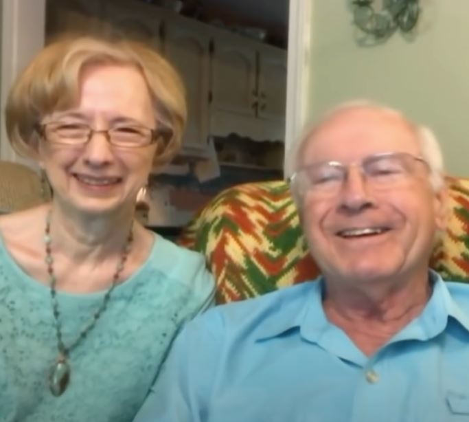Smiling elderly couple sitting closely together on a couch in a cozy home, both wearing light blue clothing and glasses.