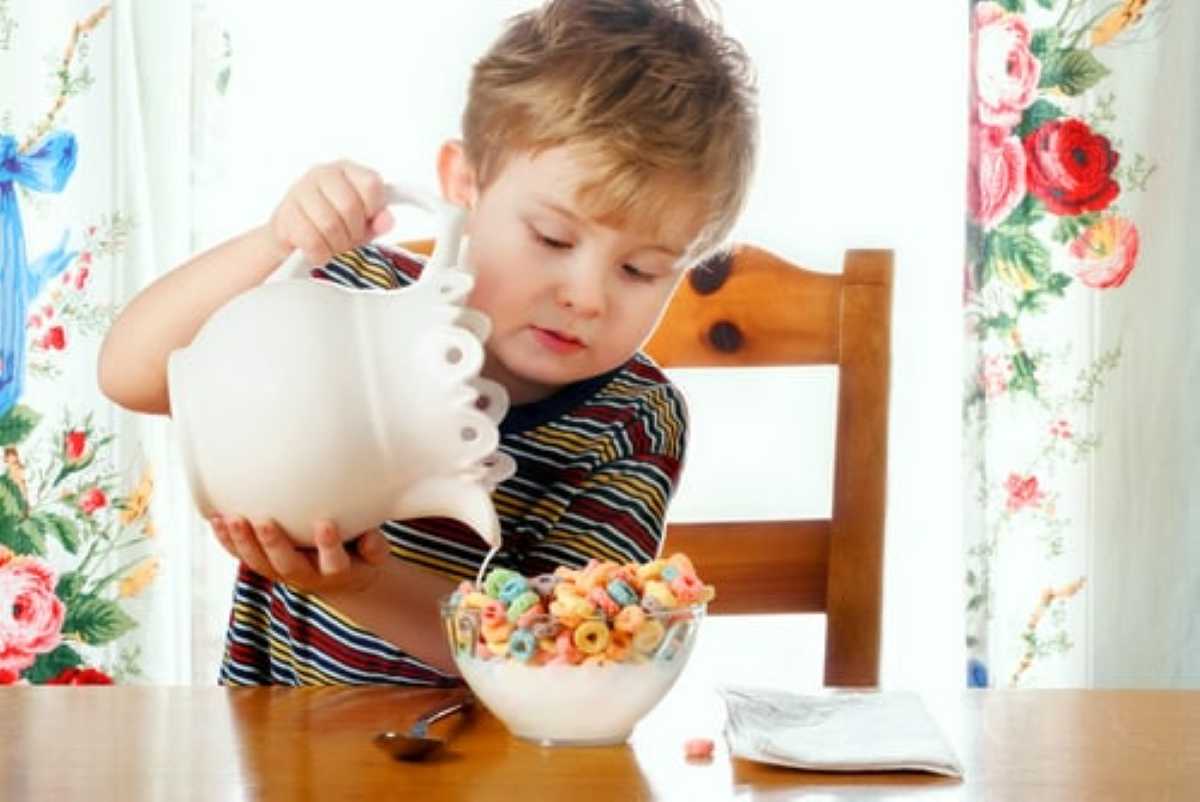 A child in a striped shirt pours milk from a white pitcher into a bowl of colorful cereal, set on a wooden table with floral curtains.