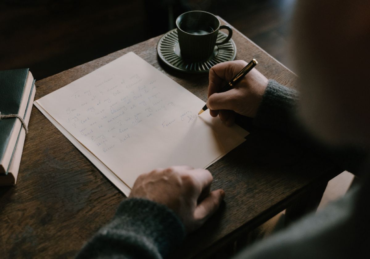 A person writes a heartfelt letter on cream paper while holding a pen, with a cup of coffee on a saucer nearby.
