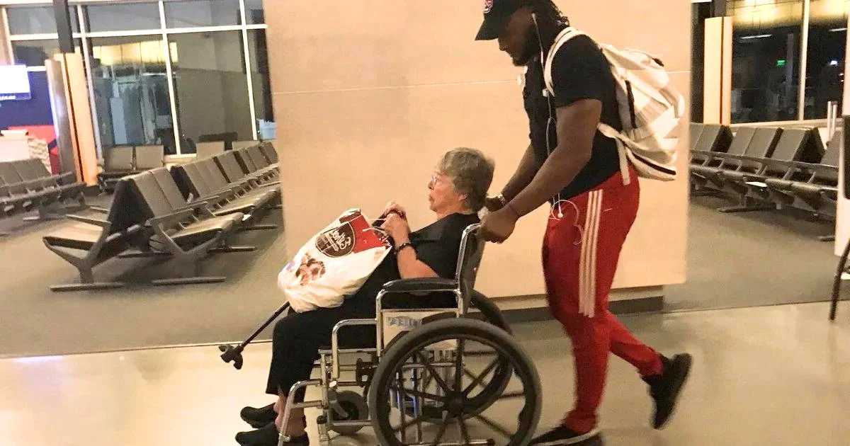 Young man pushing an elderly woman in a wheelchair through an airport terminal with empty seats in the background.