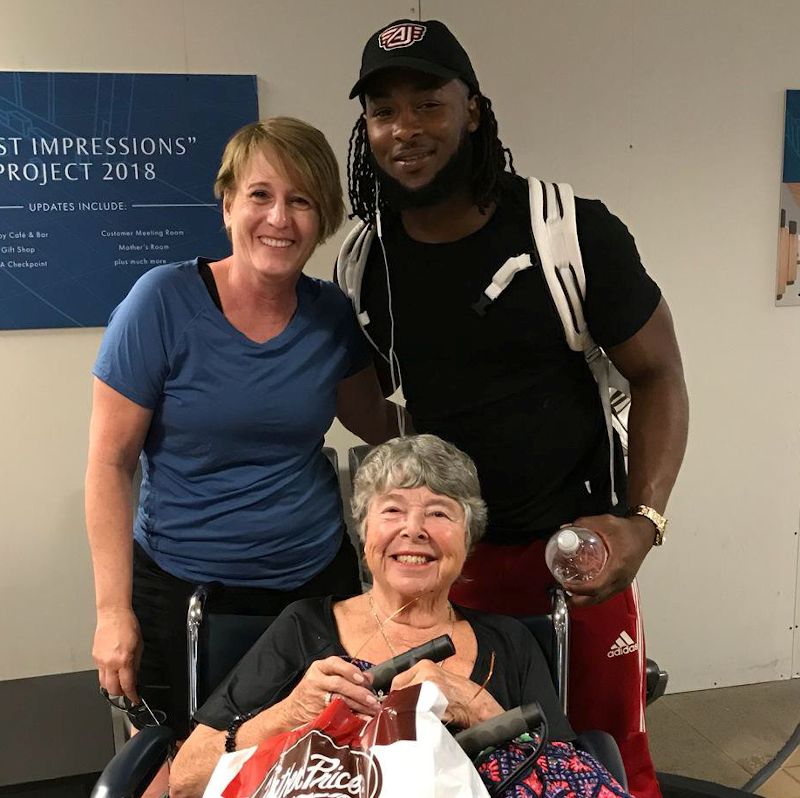 Smiling elderly woman in a wheelchair poses with a young man and another woman inside an airport near a "First Impressions Project" sign.