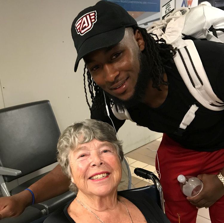 Young man kneels beside a smiling elderly woman in a wheelchair, both posing for a close-up photo inside an airport.