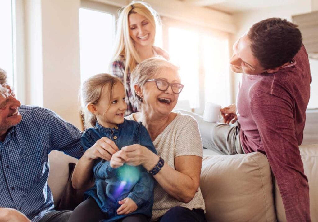 Multi-generational family laughing together on a couch in a bright living room, with a grandmother holding her smiling granddaughter.