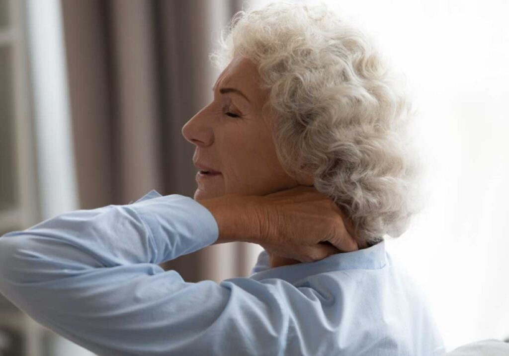 Senior woman with gray curly hair touching her neck, appearing to experience neck pain or discomfort, in a softly lit room.