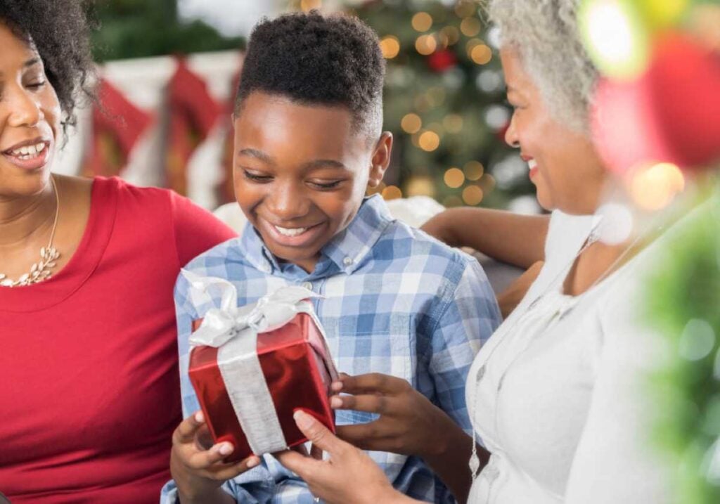 Smiling boy opening a red and silver gift box while sitting between two women, with a decorated Christmas tree in the background.