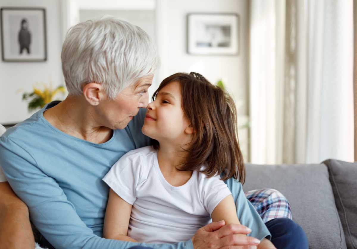 Elderly woman and young girl sitting close on a couch, touching noses and smiling affectionately in a cozy, sunlit living room.