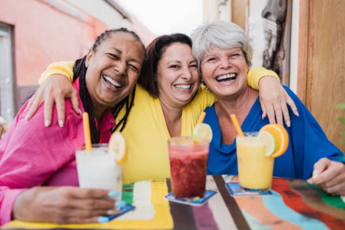 Three friends share a joyful moment at a colorful table, holding vibrant cocktails with fresh fruit garnishes.