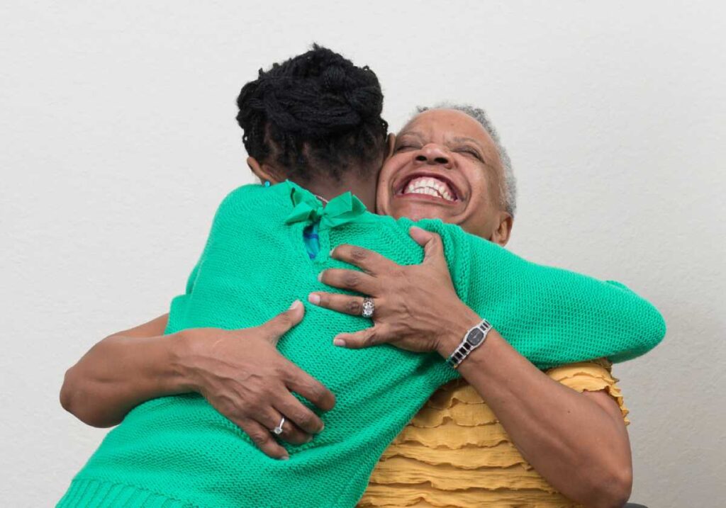 Two women share a joyful hug, one smiling widely while embracing the other who wears a bright green sweater.