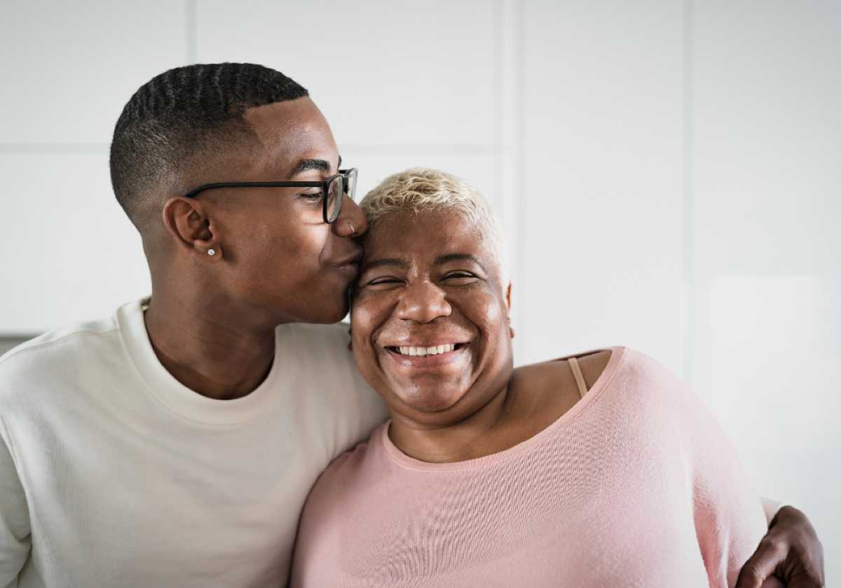 Young man kisses a smiling older woman on the cheek in a bright, modern kitchen.