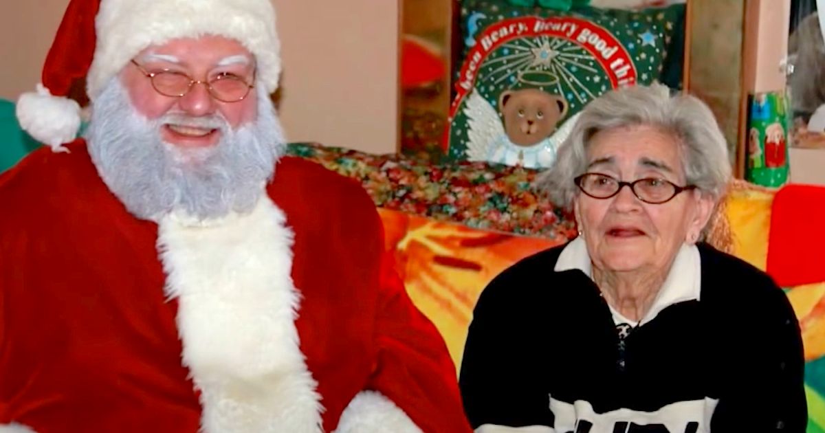 An elderly woman in a black sweater sits next to a Santa Claus figure in a cozy, festive setting, surrounded by colorful decorations.