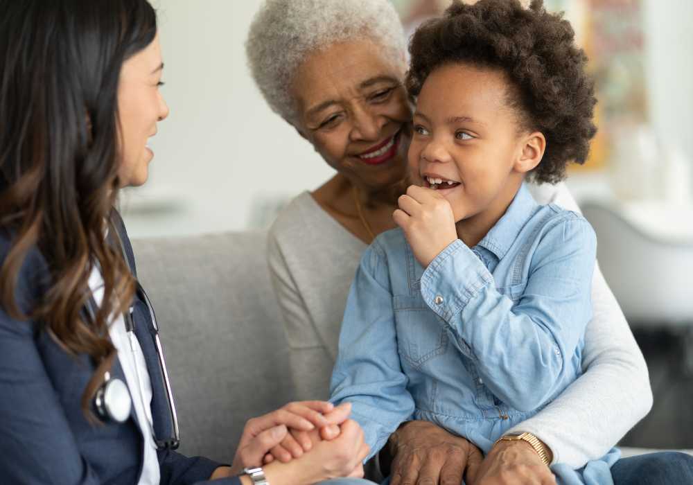 Grandmother holds young boy while they talk with a smiling female doctor during a checkup.