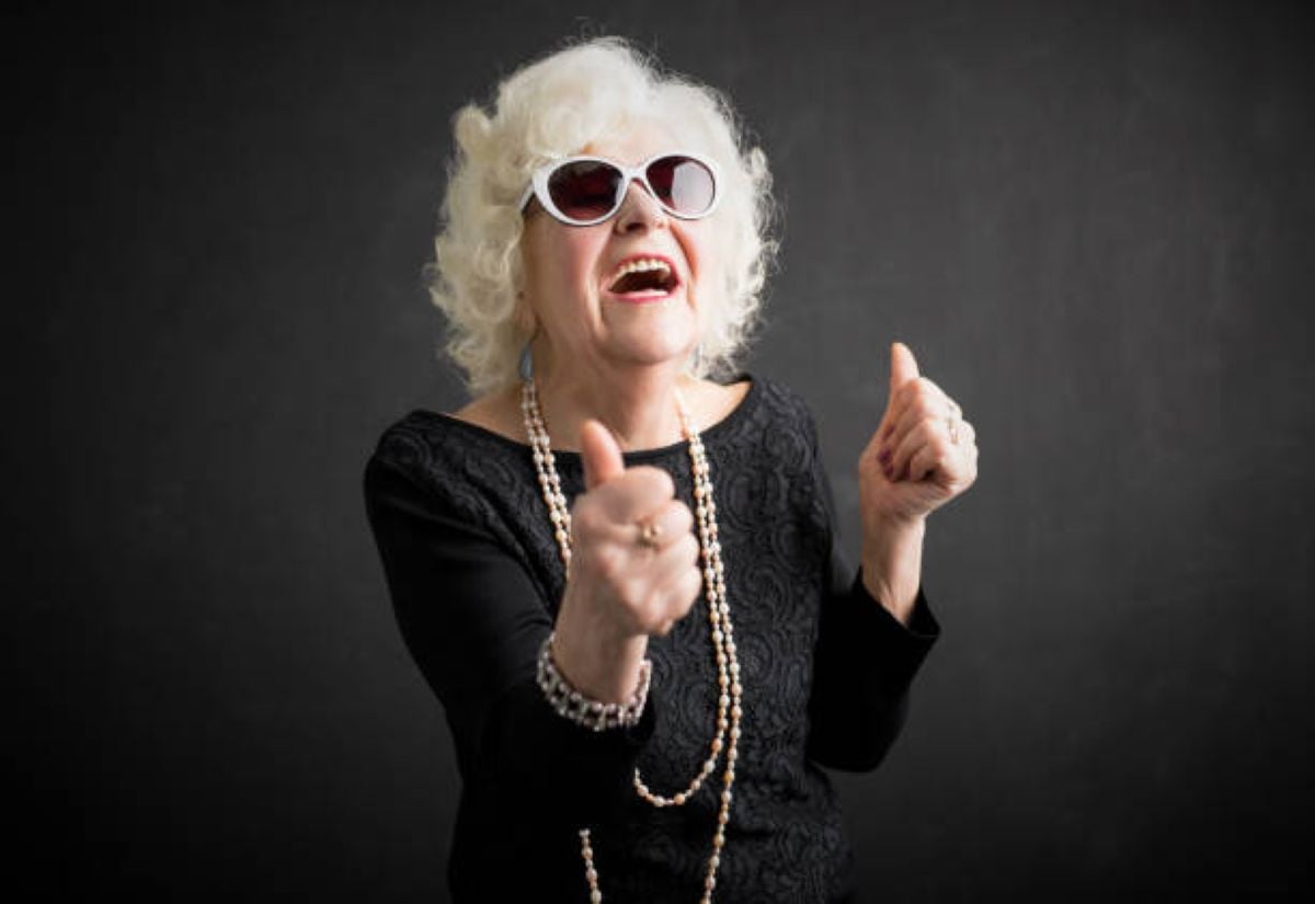 Stylish elderly woman with curly white hair wearing sunglasses and a black outfit, smiling and giving two thumbs up in front of a dark background.