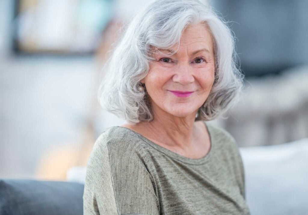 Graceful older woman with gray hair and a warm smile, sitting indoors and looking directly at the camera.