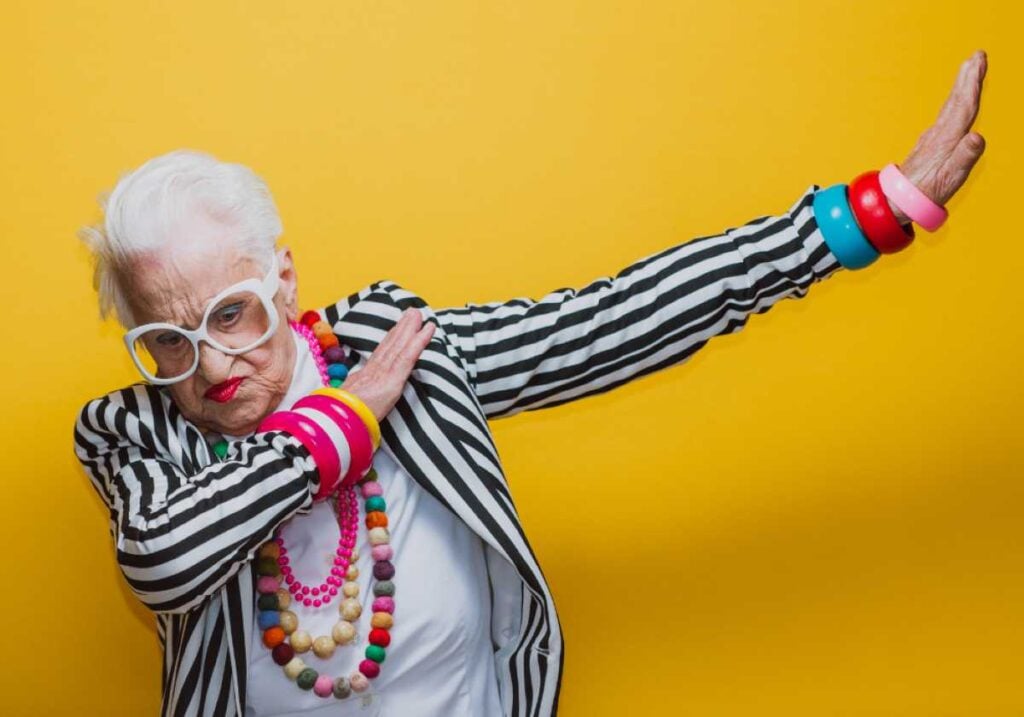 Energetic elderly woman in bold accessories and a black-and-white striped jacket striking a playful dab pose against a bright yellow background.
