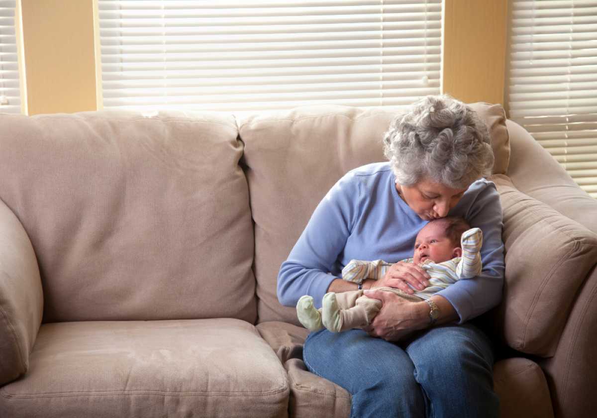 An elderly woman holds a swaddled baby on a beige couch, bathed in soft natural light from nearby windows.