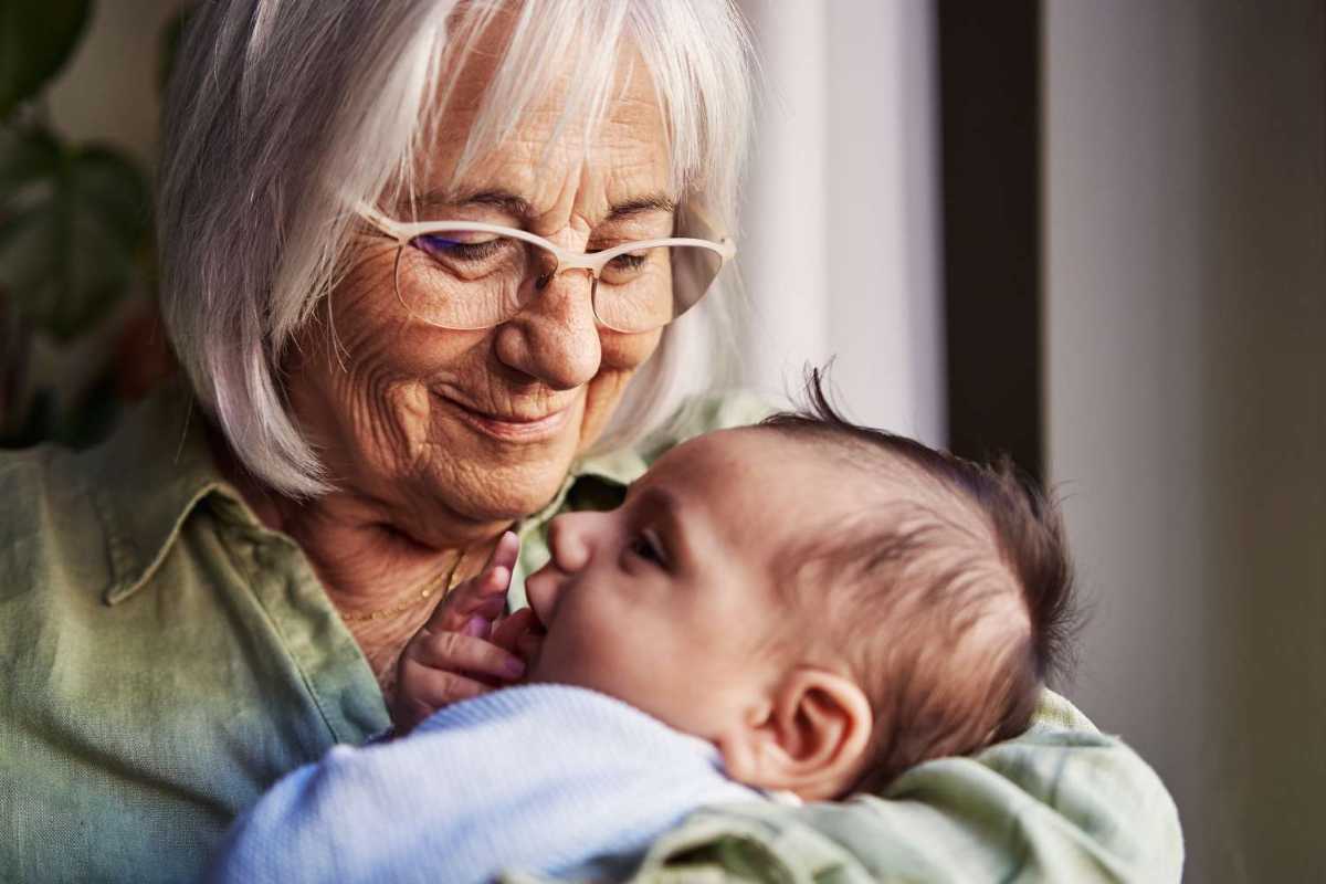 Senior woman with glasses gazing tenderly at a baby in her arms, bathed in soft natural light near a window.