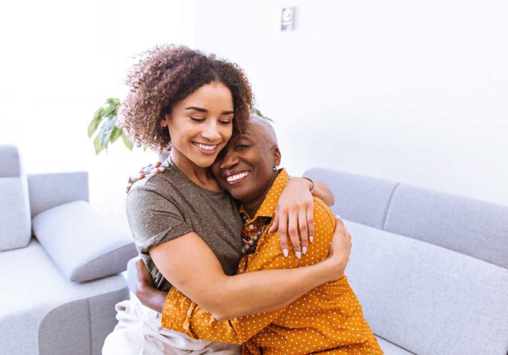 Two women share a warm embrace on a couch, smiling with eyes closed in a bright, cozy living room.