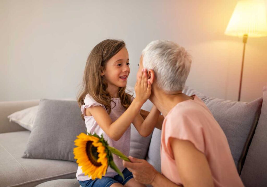 Young girl holding a sunflower gently touches an older woman’s face as they smile at each other on a couch.