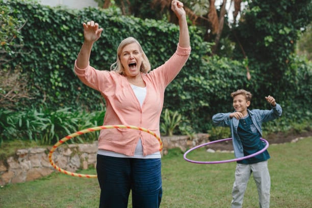 A woman and a child are playing with hula hoops in a sunny backyard, surrounded by greenery and a stone wall.