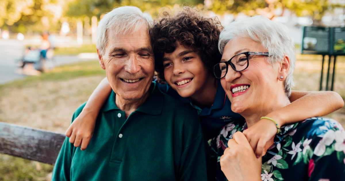 A child stands between two smiling adults, embracing them as they sit on a park bench surrounded by greenery.