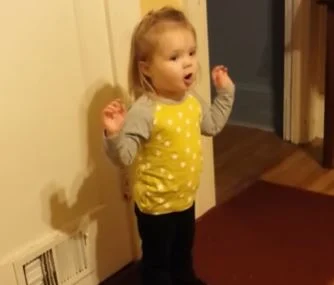 Little girl in a yellow polka dot shirt standing indoors with her hands raised, looking surprised or mid-sentence.