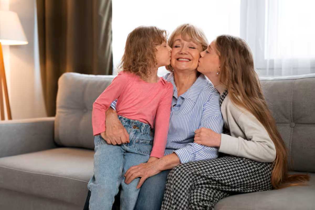 Smiling grandmother gets kissed on both cheeks by two young girls while sitting on a couch.