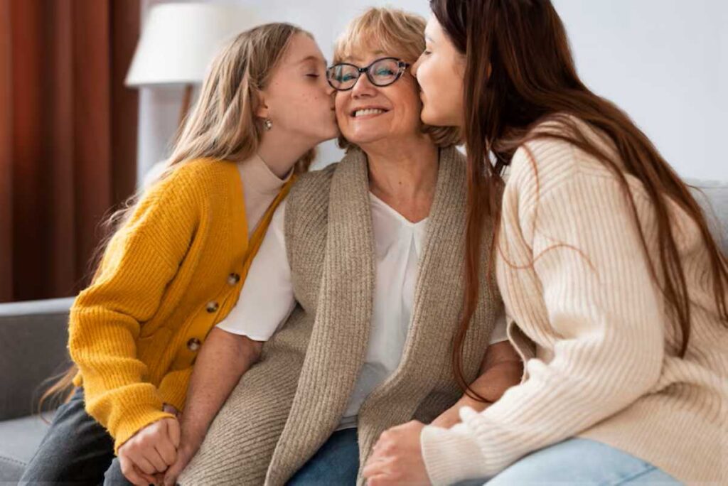 Grandmother in glasses is kissed on the cheeks by two young women in cozy sweaters.