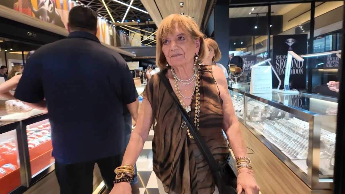 Older woman wearing layered jewelry walks through a brightly lit jewelry store, surrounded by glass display cases and other shoppers.