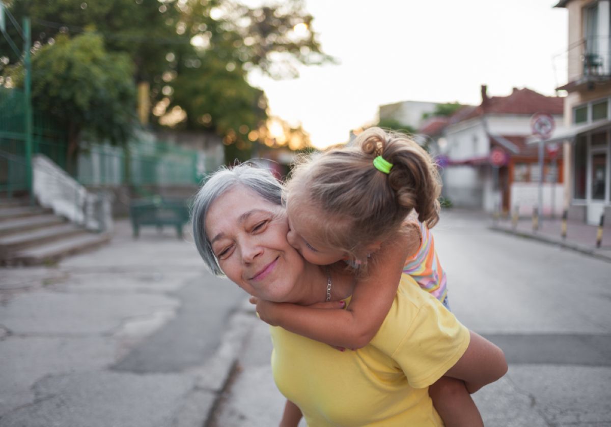 Smiling older woman getting a kiss on the cheek from a young girl riding on her back outdoors in a quiet neighborhood.
