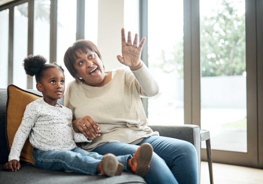 Grandmother and granddaughter sitting on a couch, holding hands and waving happily while looking toward something outside.