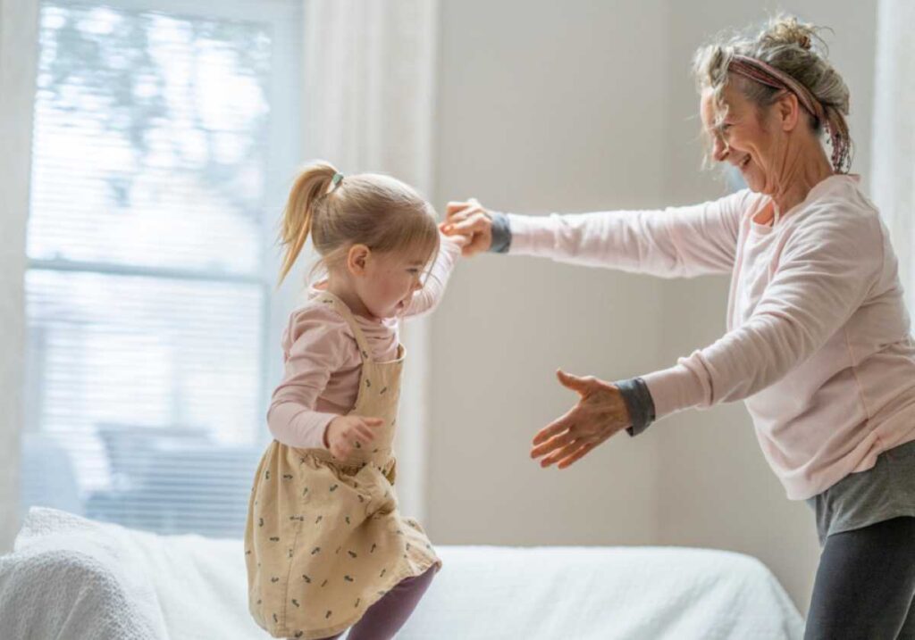 Joyful grandmother catching her young granddaughter mid-jump on a couch in a bright, cozy living room.