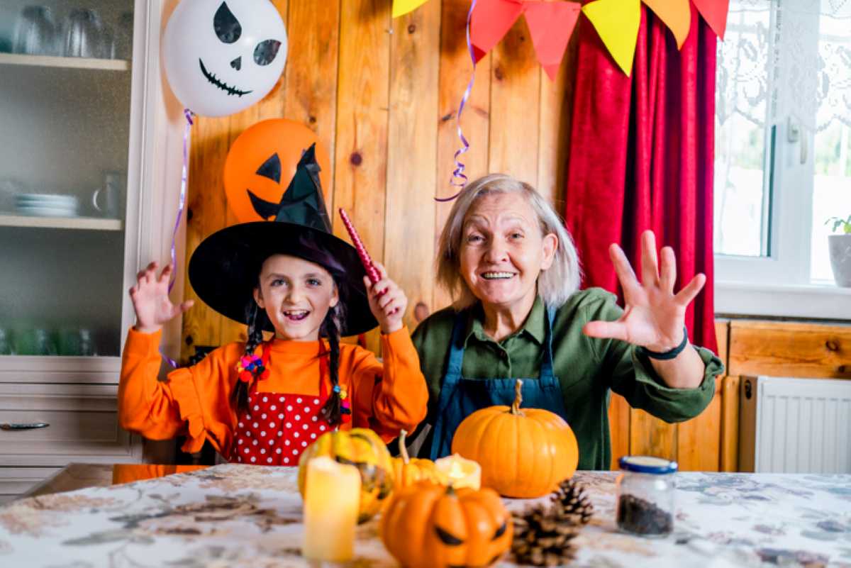 Smiling girl in a witch costume and older woman wave at the camera while sitting at a table decorated with pumpkins and Halloween decor.
