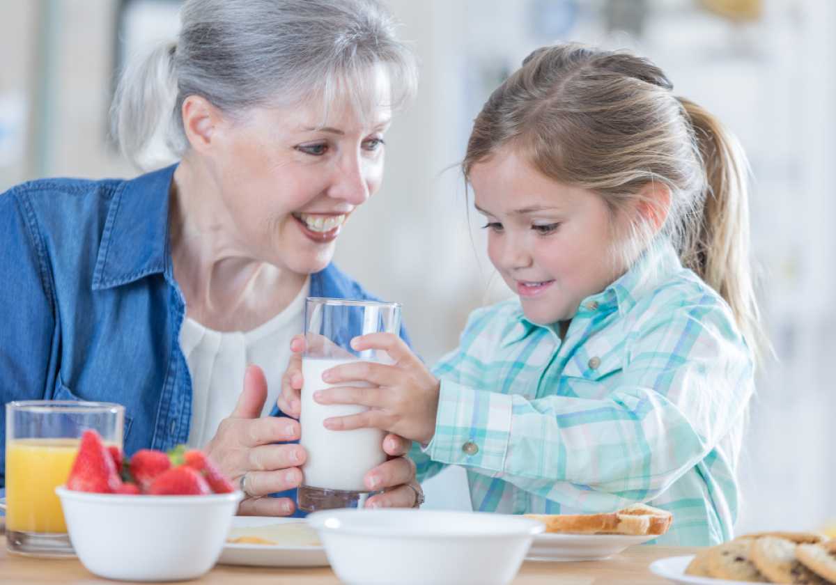 Smiling grandmother and young girl sharing a glass of milk at the breakfast table with fruit, juice, and toast.